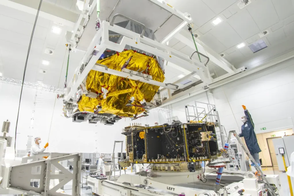 La salle blanche où des ingénieurs travaillent sur l'assemblage des composants de vaisseau spatial. Un grand module enveloppé de feuille dorée est délicatement descendu par une grue sur une autre section d'un vaisseau spatial positionnée en dessous. La scène comprend des personnes en combinaisons de salle blanche, des équipements de levage spécialisés et divers appareils techniques, soulignant la précision et le soin requis dans l'ingénierie aérospatiale.