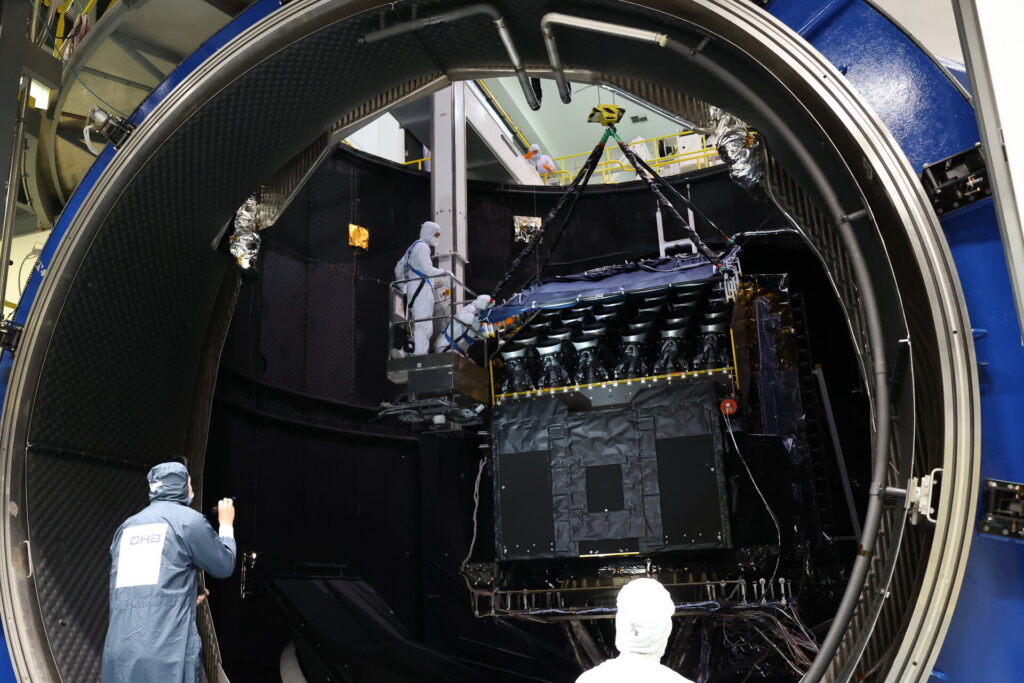 Le satellite PLATO au centre de l'image placé dans la chambre à vide de l'EA. Il est en train d'être levé via des sangles pour sortir de la chambre à vide. Des ingénieurs en tenus propre aide et regarde le processus pour assurer la sortie en sécurité.