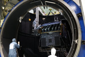 Le satellite PLATO au centre de l'image placé dans la chambre à vide de l'EA. Il est en train d'être levé via des sangles pour sortir de la chambre à vide. Des ingénieurs en tenus propre aide et regarde le processus pour assurer la sortie en sécurité.