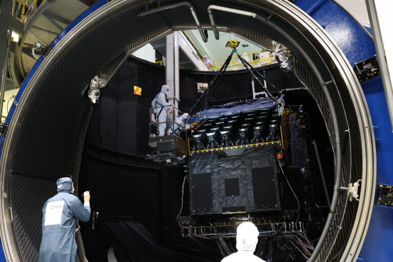 Le satellite PLATO au centre de l'image placé dans la chambre à vide de l'EA. Il est en train d'être levé via des sangles pour sortir de la chambre à vide. Des ingénieurs en tenus propre aide et regarde le processus pour assurer la sortie en sécurité.