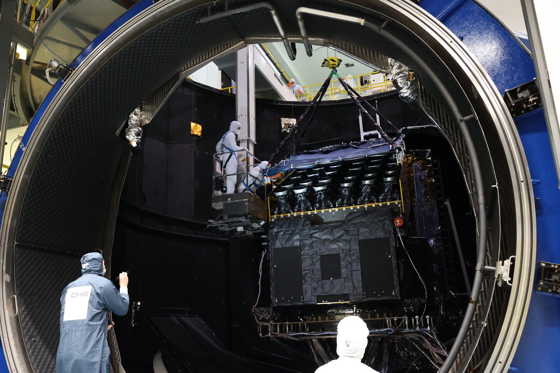 Le satellite PLATO au centre de l'image placé dans la chambre à vide de l'EA. Il est en train d'être levé via des sangles pour sortir de la chambre à vide. Des ingénieurs en tenus propre aide et regarde le processus pour assurer la sortie en sécurité.